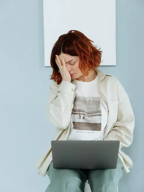 ruda kobieta siedzi z laptopem na kolanach z ręką przy twarzy, wygląda na załamaną i wyczerpaną